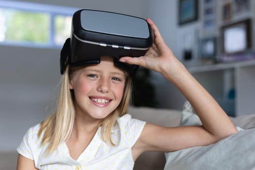 Portrait of caucasian girl sitting on couch wearing vr headset raising it and smiling. staying at home in isolation during quarantine lockdown.