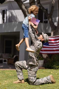 Smiling caucasian soldier father lifting happy son in garden with american flag outside house. soldier returning home to family.
