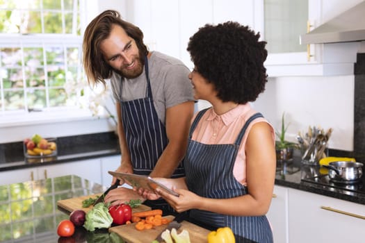 Happy diverse couple in kitchen using tablet and preparing food. staying at home in isolation during quarantine lockdown.