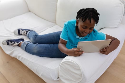 Smiling african american boy with short dreadlocks lying on couch using digital tablet. staying at home in isolation during quarantine lockdown.