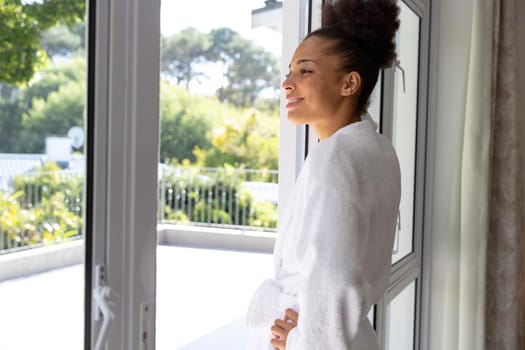 Mixed race woman wearing bathrobe standing by terrace door and smiling. staying at home in isolation during quarantine lockdown.