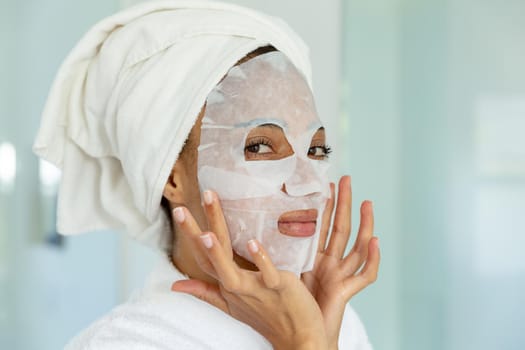 Mixed race woman wearing bathrobe and cleansing face mask in bathroom. staying at home in isolation during quarantine lockdown.