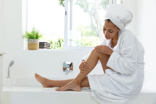 Mixed race woman wearing bathrobe and putting cream on her legs in bathroom. staying at home in isolation during quarantine lockdown.