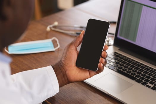 Mid section of african american senior male doctor holding smartphone with copy space at home. distant communication and telemedicine consultation concept.