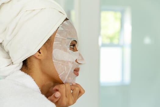 Mixed race woman wearing bathrobe and cleansing face mask in bathroom. staying at home in isolation during quarantine lockdown.