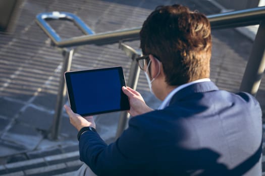 Asian businessman wearing face mask using tablet sitting on steps in city street. digital nomad out and about in city during covid 19 pandemic concept.