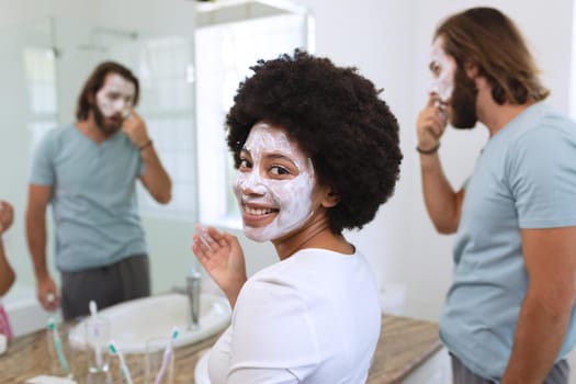 Portrait of happy diverse couple standing in bathroom wearing beauty masks. staying at home in isolation during quarantine lockdown.