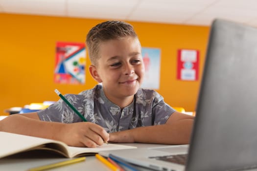 Caucasian boy sitting at a desk in classroom writing, looking at laptop and smiling. childhood, technology and education at elementary school.