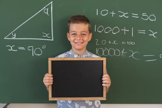 Portrait of caucasian boy at chalkboard with geometry on it, holding blank chalkboard, smiling. childhood and education at elementary school.