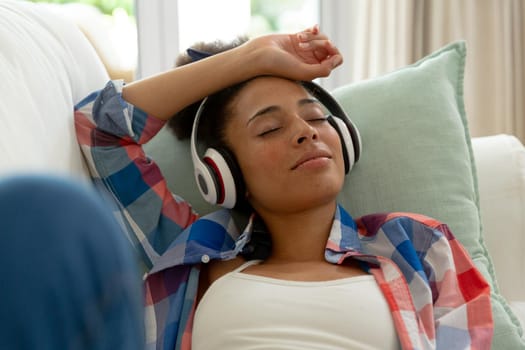 Mixed race woman lying on couch listening to music with headphones at home. staying at home in isolation during quarantine lockdown.