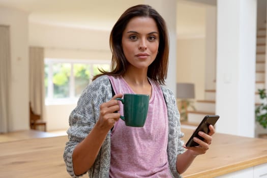 Caucasian woman standing in kitchen drinking cup of coffee using smartphone and smiling. staying at home in isolation during quarantine lockdown.