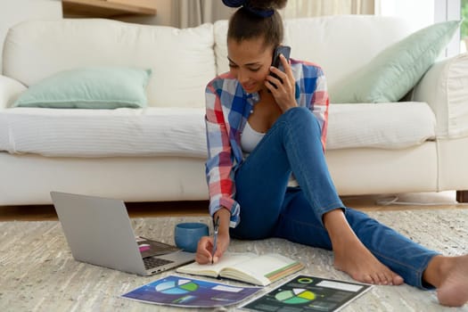 Mixed race woman sitting on the floor working from home using laptop and smartphone. staying at home in isolation during quarantine lockdown.
