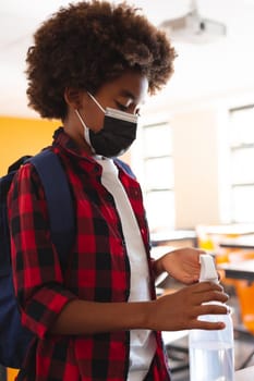 African american schoolboy in face mask standing in classroom disinfecting hands. childhood and education at elementary school during coronavirus covid19 pandemic.