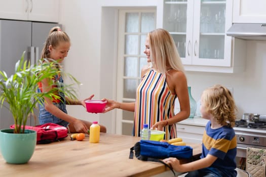 Smiling caucasian mother with son and daughter preparing packed lunches and school bags in kitchen. happy family spending time together at home.