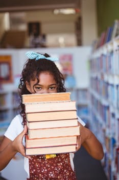 Portrait of happy african american schoolgirl carrying stack of books in school library. childhood and education at elementary school.