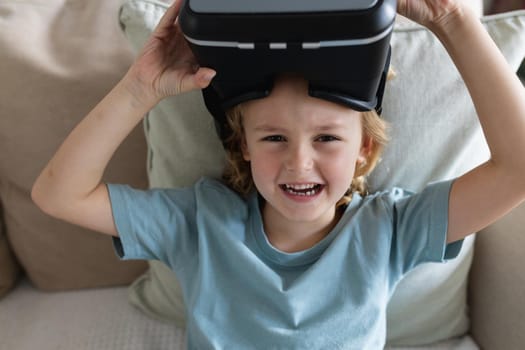 Portrait of caucasian boy sitting on couch wearing vr headset raising it and laughing. staying at home in isolation during quarantine lockdown.