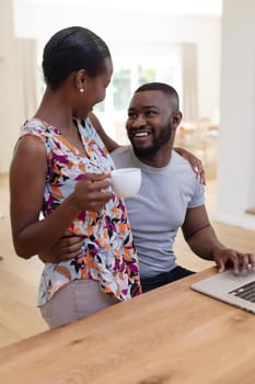 Happy african american couple embracing in kitchen, man using laptop woman holding coffee. staying at home in isolation during quarantine lockdown.