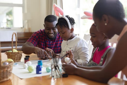African american parents with son and daughter wearing bunny ears painting colourful eggs. celebrating easter at home in isolation during quarantine lockdown.