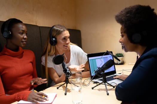 Diverse group of female business colleagues wearing headphones in discussion speaking to microphones. recording podcast and business in a modern office.