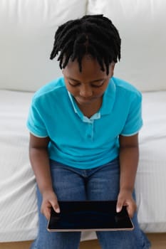 High angle view of african american boy with short dreadlocks sitting on couch using digital tablet. staying at home in isolation during quarantine lockdown.