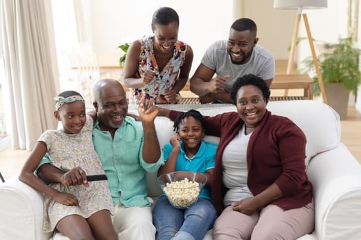 Excited african american parents, grandparents and grandchildren on couch watching tv and cheering. happy three generation family spending time together at home.