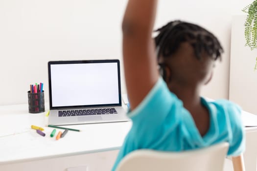 African american boy at desk using laptop with copy space for online school lesson and raising hand. staying at home in isolation during quarantine lockdown.