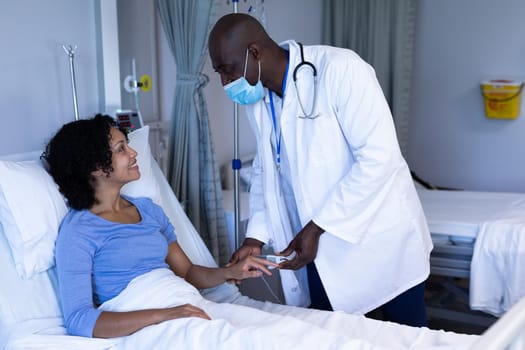 African american male doctor in face mask puts oximeter on finger of female patient in hospital bed. medicine, health and healthcare services during coronavirus covid 19 pandemic.