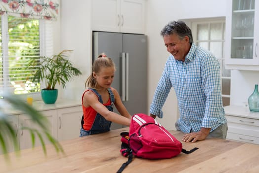 Caucasian grandfather in kitchen with granddaughter packing rucksack. happy family spending time together at home.