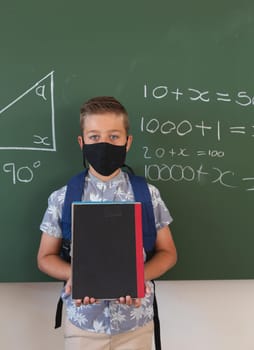 Portrait of caucasian boy in face mask standing at chalkboard with geometry on it holding books. childhood and education at elementary school during coronavirus covid19 pandemic.
