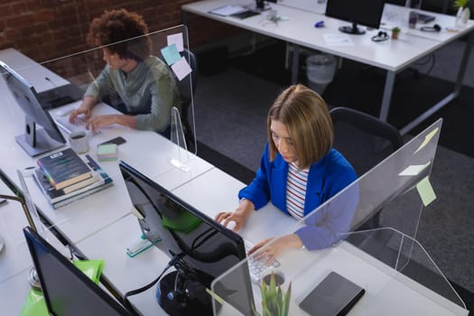 Diverse male and female colleagues sitting in front of computers separated by sneeze shields. independent creative design business during covid 19 coronavirus pandemic.