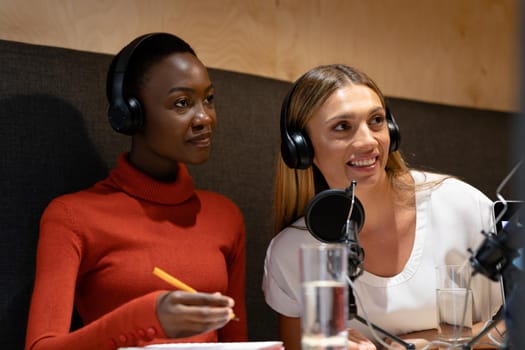 Diverse female business colleagues wearing headphones in discussion speaking to microphones. recording podcast and business in a modern office.