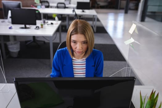 Asian businesswoman sitting in office in front of computer near sneeze shield. independent creative design business during covid 19 coronavirus pandemic.