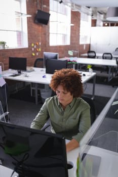 Mixed race businessman sitting in office in front of computer near sneeze shield. independent creative design business during covid 19 coronavirus pandemic.