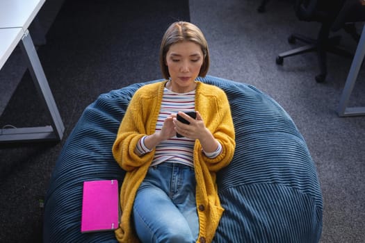 Asian businesswoman chilling in office relaxing space lying in beanbag looking at smartphone. independent creative design business.