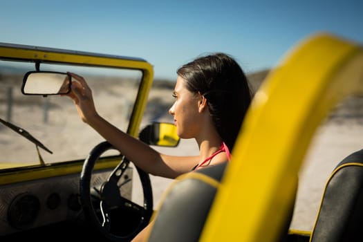 Happy caucasian woman sitting in beach buggy by the sea looking at herself in car mirror. beach break on summer holiday road trip.