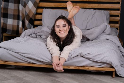 Skinny young woman stretching in bed feeling truly relaxed. pretty girl chilling wearing knitted beige sweater in bedroom at home.