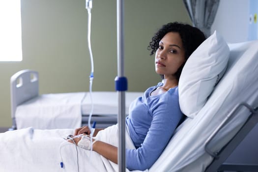 Mixed race female patient sitting in hospital bed wearing fingertip pulse oximeter and iv drip. medicine, health and healthcare services.