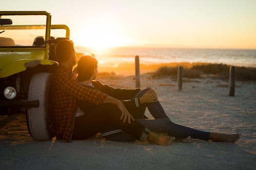 Happy caucasian couple leaning against beach buggy by the sea watching sunset. beach break on summer holiday road trip.