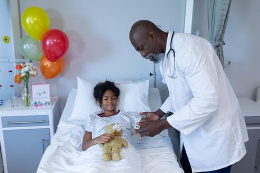 African american male doctor putting pulse oximeter on sick mixed race girl in hospital bed. medicine, health and healthcare services.