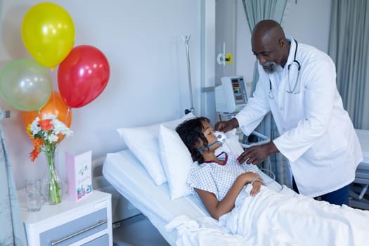 African american male doctor putting oxygen mask ventilator on sick mixed race girl in hospital bed. medicine, health and healthcare services during coronavirus covid 19 pandemic.