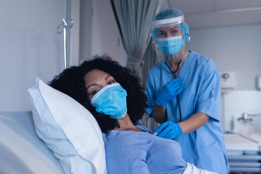 Caucasian female doctor in hospital in face mask examining female patient with stethoscope. medical professional at work during coronavirus covid 19 pandemic.