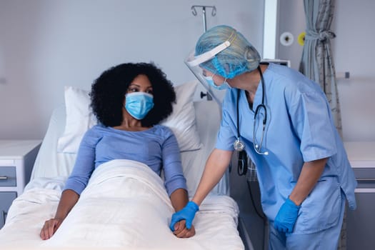 Caucasian female doctor in hospital in face mask consoling female patient in bed wearing face mask. medical professional at work during coronavirus covid 19 pandemic.