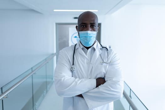 Portrait of african american male doctor in hospital corridor with arms crossed wearing face mask. medical professional at work during coronavirus covid 19 pandemic.