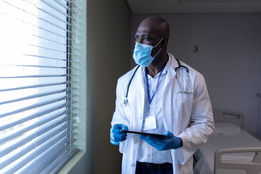 African american male doctor in hospital wearing face mask using tablet. medical professional at work during coronavirus covid 19 pandemic.