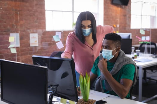 Diverse business colleagues wearing face masks brainstorming in front of computer. independent creative design business during covid 19 coronavirus pandemic.