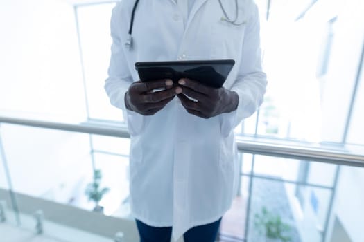 Midsection of african american male doctor in hospital corridor using tablet. medical professional at work, medicine and healthcare.