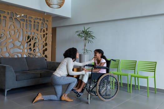 Happy mixed race mother kneeling holding hand of smiling daughter in wheelchair in hospital foyer. medicine, health and healthcare services.