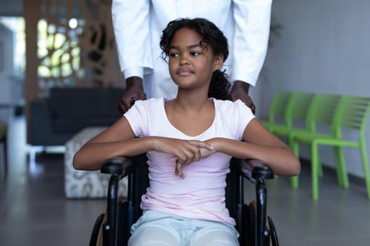 African american male doctor pushing smiling mixed race girl in wheelchair through hospital corridor. medicine, health and healthcare services.