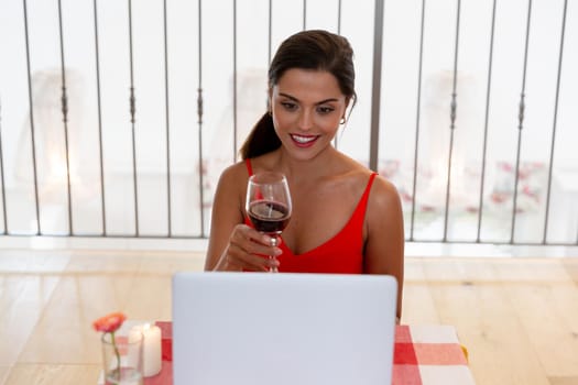 Caucasian woman sitting by desk having a video chat on laptop drinking wine and smiling. staying at home in isolation during quarantine lockdown.