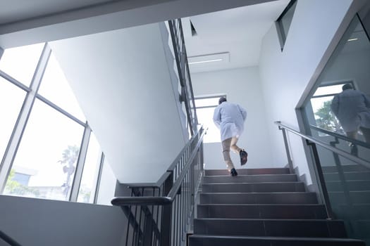Senior african american male doctor wearing lab coat running up stairs in hospital. medical professional at work.